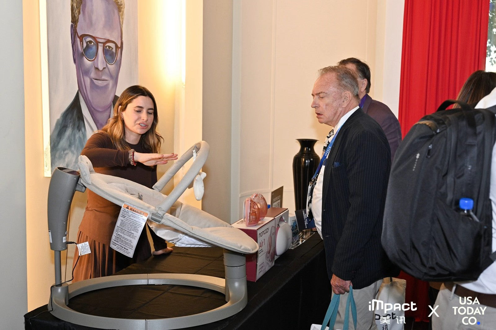 Nicolas Charbonnier A woman gesturing towards the Momcozy 2-in-1 Electric Baby Swing while explaining its features to a man in a suit at an exhibition booth