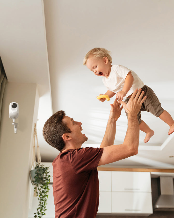 Father lifting laughing baby in modern kitchen, with 4.3-inch HD video baby monitor mounted on wall in the background.