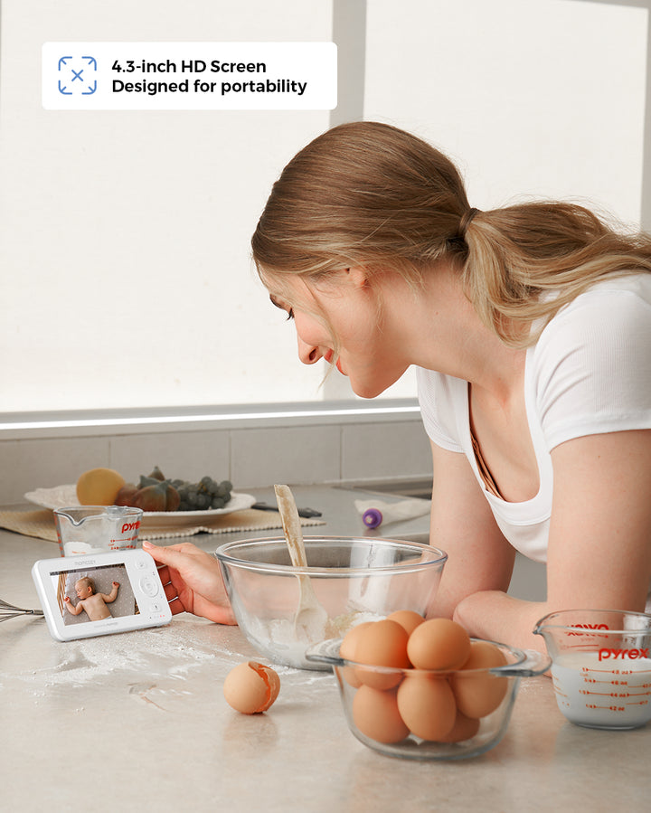 Woman in kitchen looking at 4.3-inch HD video baby monitor displaying a baby. Kitchen counter with glass bowl, whisk, eggs, measuring cup, and baking ingredients. Text reads '4.3-inch HD Screen Designed for portability.'