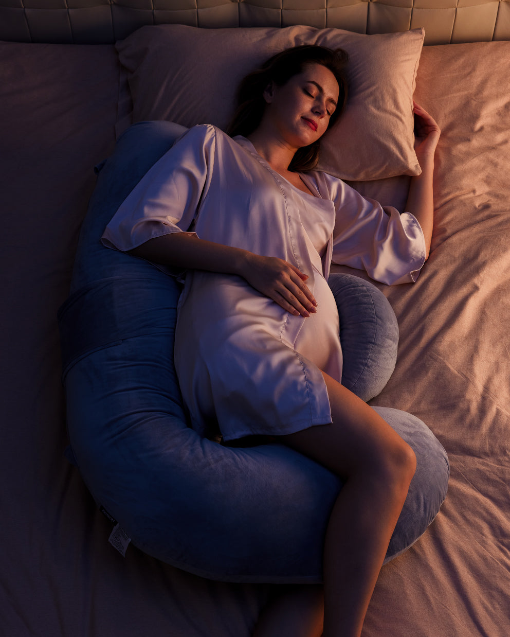 Pregnant woman sleeping soundly on a gray F-shaped pregnancy pillow for full body support during pregnancy. Momcozy pillow.