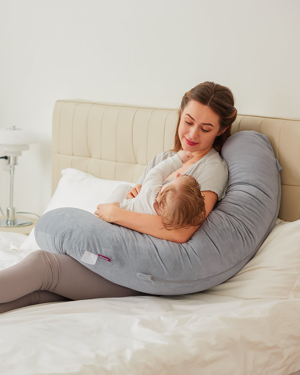 Woman with baby using gray Momcozy F-shaped pregnancy pillow for support and comfort during feeding or resting.