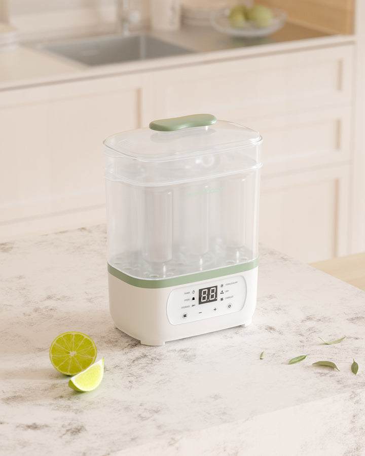 8-in-1 baby bottle steam sterilizer on a kitchen counter with digital LED countdown display, transparent lid, and green accents, next to lime slices and green leaves.