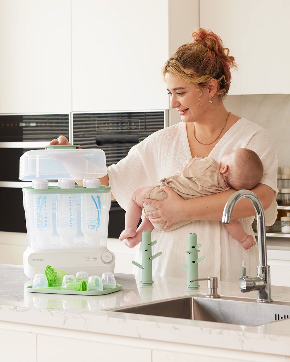 Mom holding a baby while operating Momcozy 3 Layers Bottle Sterilizer on kitchen counter.