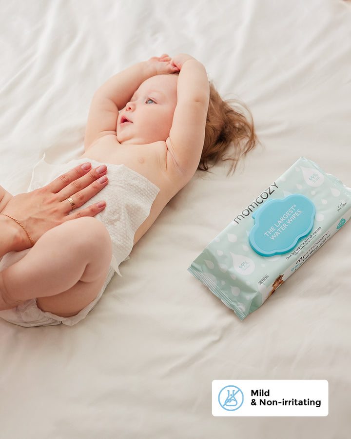 Baby lying on a white bed while an adult gently cleans with Momcozy 99% Water Wipes, featuring mild and non-irritating formula.