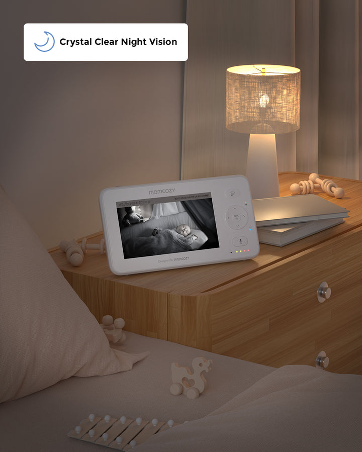 Momcozy 4.3" HD Video Baby Monitor BM02 with crystal clear night vision, showing a sleeping baby on the screen. Placed on a wooden nightstand next to a lit lamp.