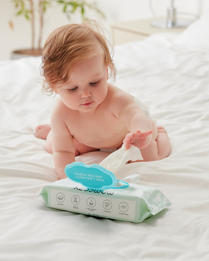 Baby reaching for a pack of Momcozy 99% Water Wipes on a bed, highlighting the product's high level of purity, hypoallergenic nature, alcohol-free content, and dermatological testing.