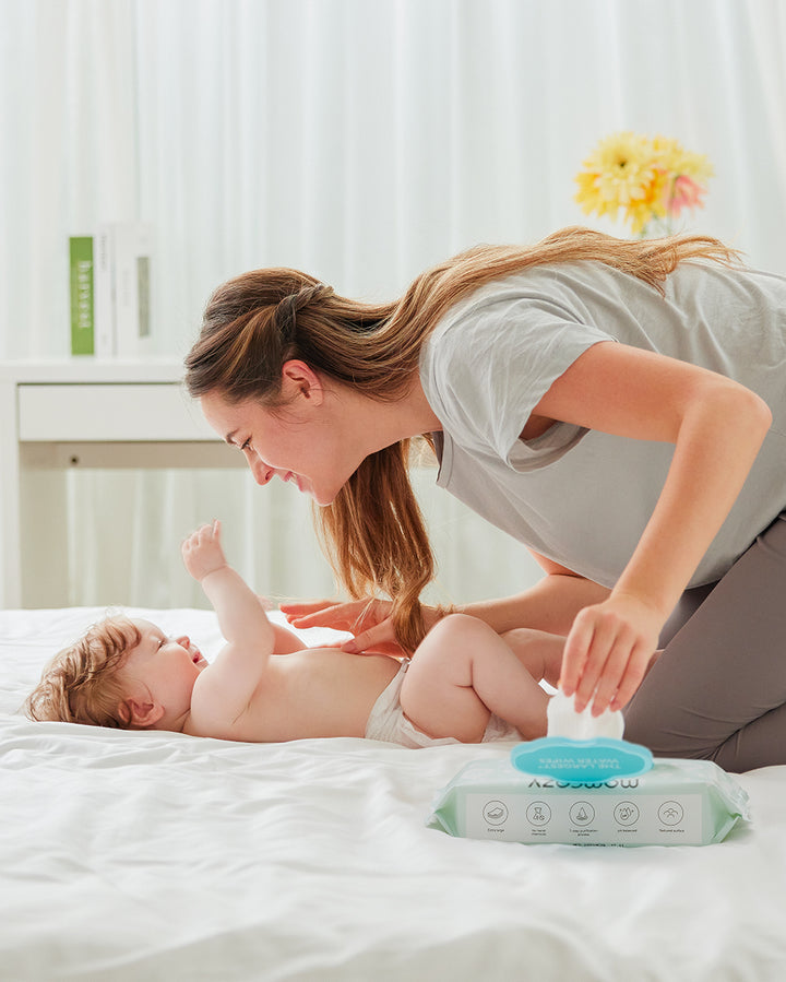 Mother changing baby's diaper using Momcozy 99% Water Wipes on a bed. Mother smiling and interacting with baby while holding a water wipe from an open package. Bright room with white curtains and yellow flower decor in the background.