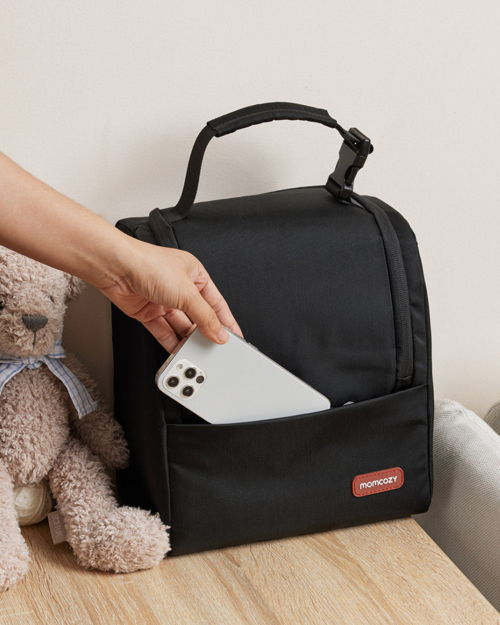 Momcozy black insulated baby bottle bag with front pocket, smartphone, and Momcozy logo.