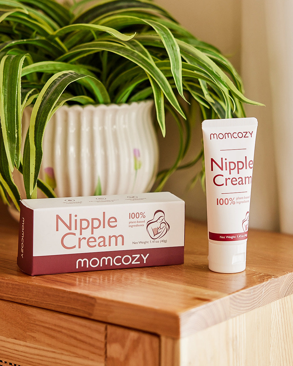 Momcozy Nipple Cream tube and box placed on a wooden surface with a green plant in the background. 100% vegan and hypoallergenic nipple cream for breastfeeding mothers.