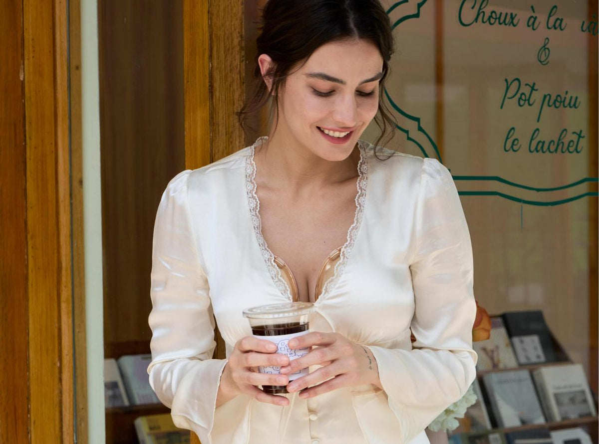 A woman wearing a cream satin long-sleeve blouse with lace trim at the neckline, holding an iced coffee in front of a shop window.